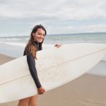 portrait-charming-pretty-woman-with-long-hair-preparing-lesson-surfing-smiles-camera-holding-surfboard-stands-s